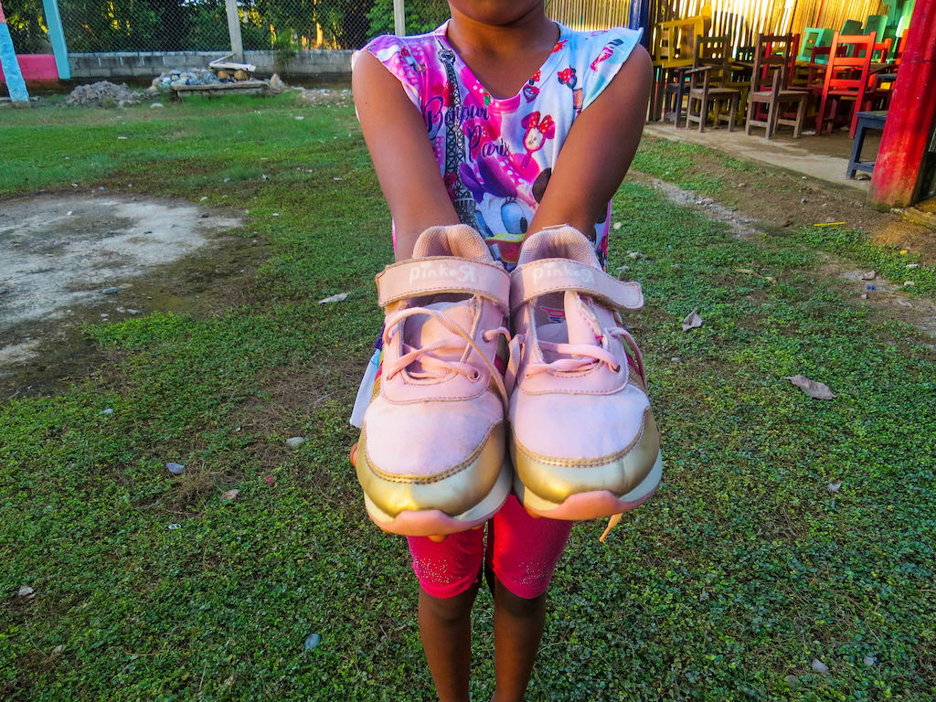 Iris is wearing a brightly coloured shirt and pink shorts. She is standing in the yard of the Compassion centre and is holding out her new shoes.