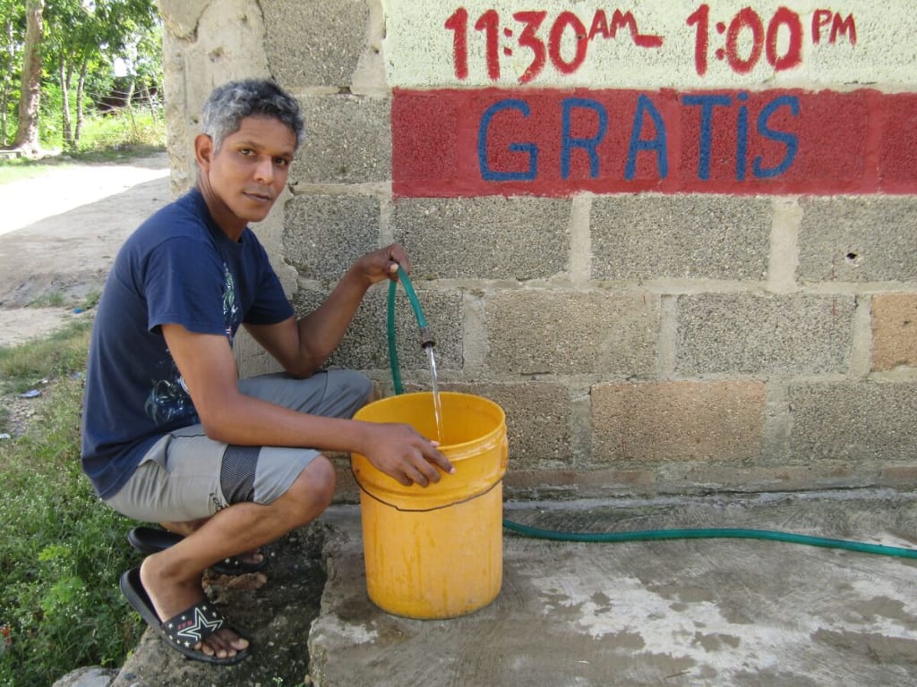 A man in a blue shirt kneels beside a yellow bucket and holds a hose.