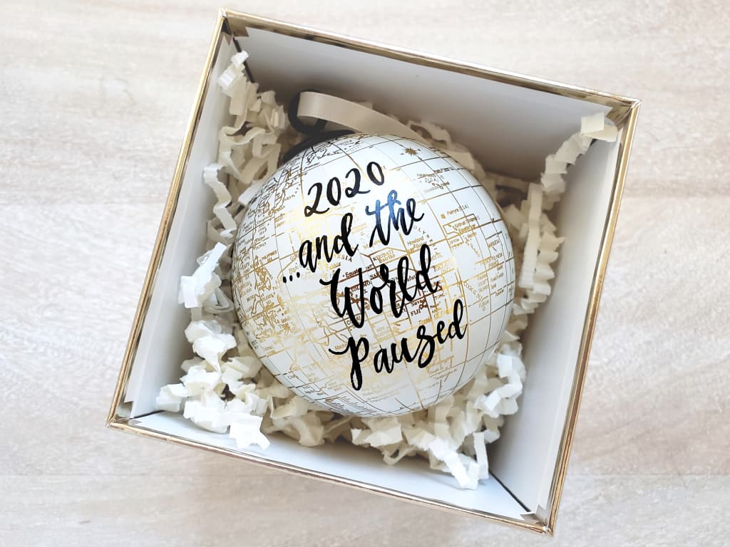 An ornament is in a box. The ornament looks like a globe with the words, "2020...and the world paused" on it.