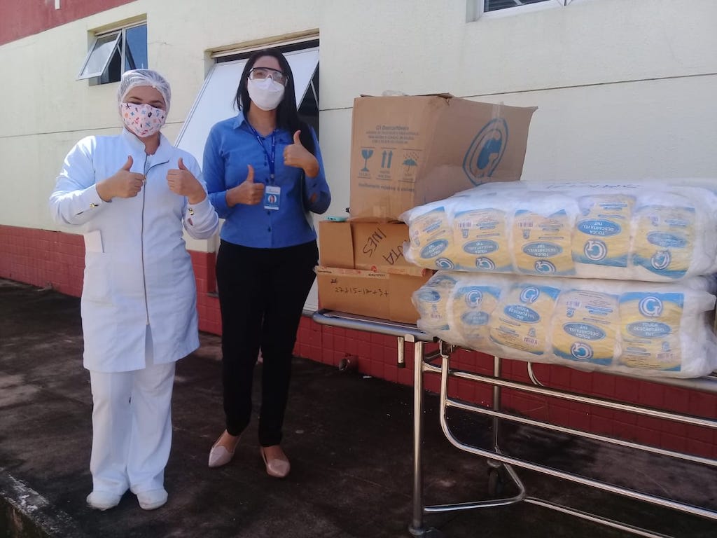 A Compassion staff member and a nurse stand together beside a table outside the hospital that is loaded with donated supplies. Both wear masks and are making a "thumbs up" sign with their hands.