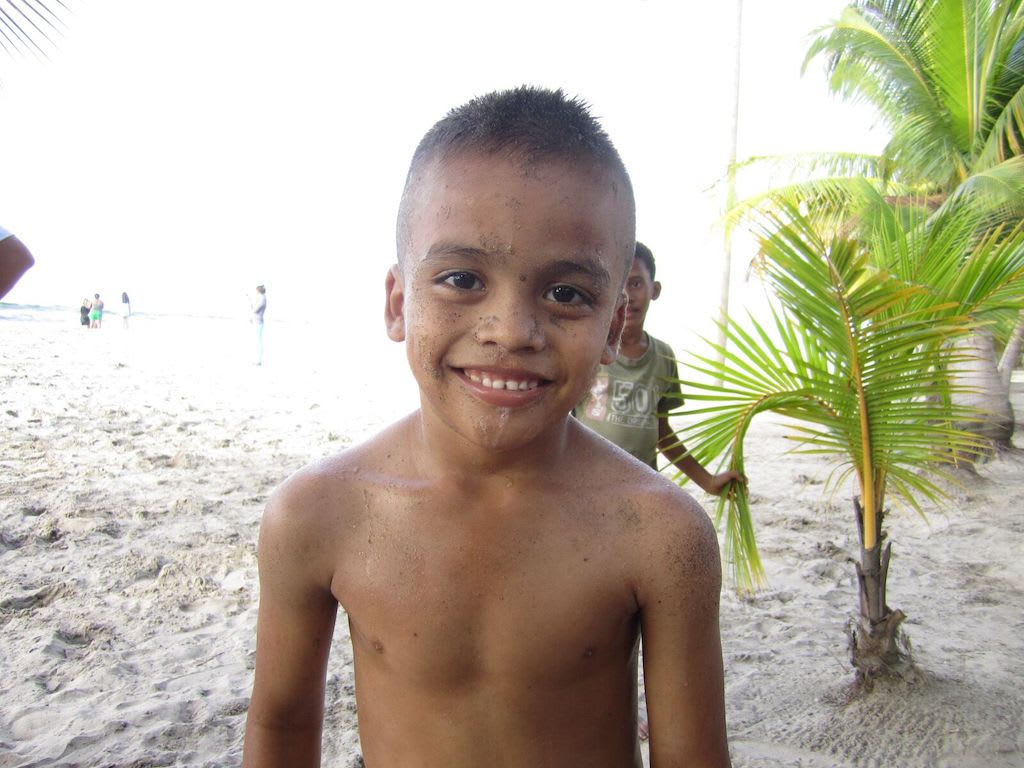 A shirtless boy covered in sand smiles at the camera on the beach.