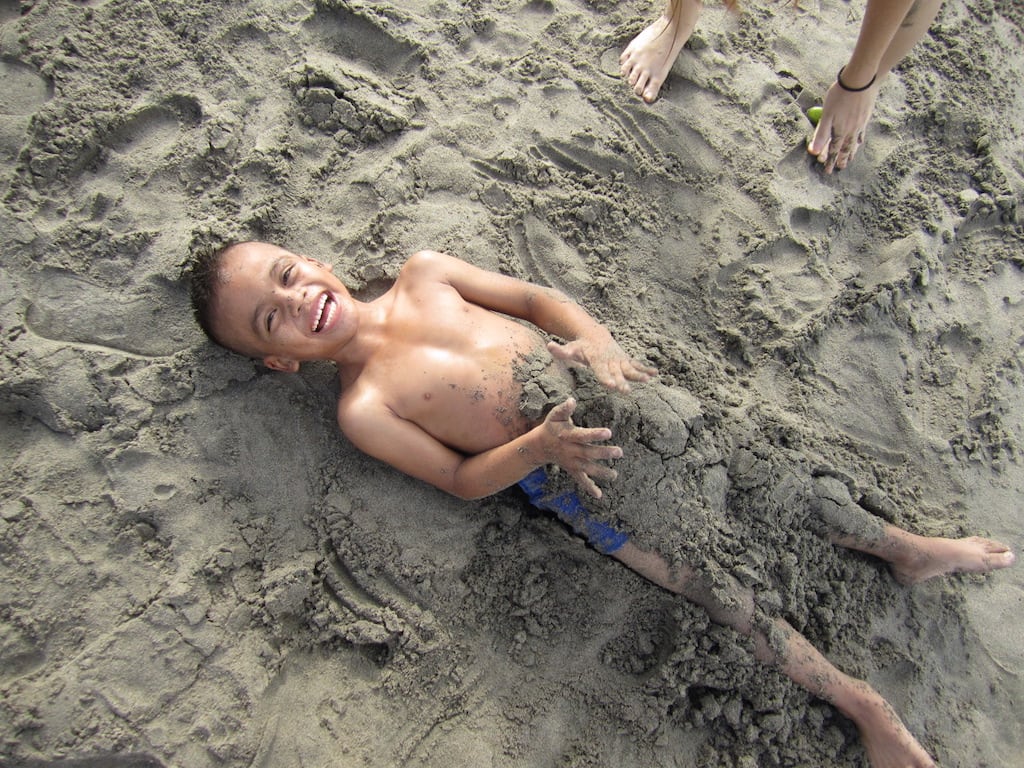 A boy lays on the beach, his legs buried in sand.