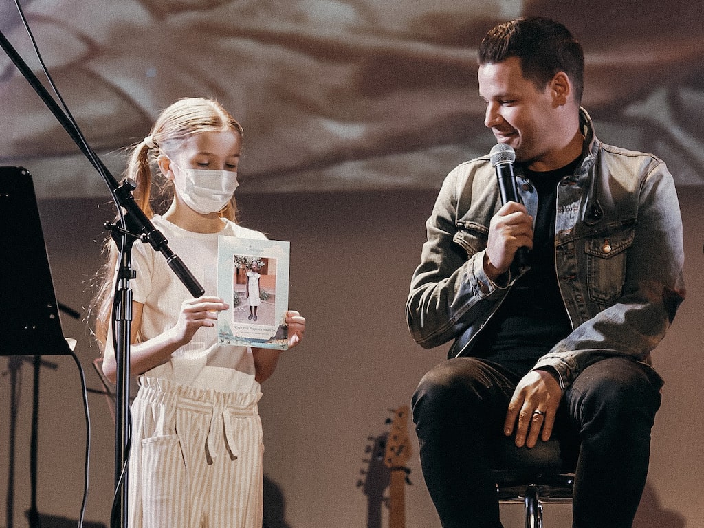 Pastor Drew sits on a stool on stage next to his daughter who is standing and holding up a photo of their family's Compassion sponsored child.