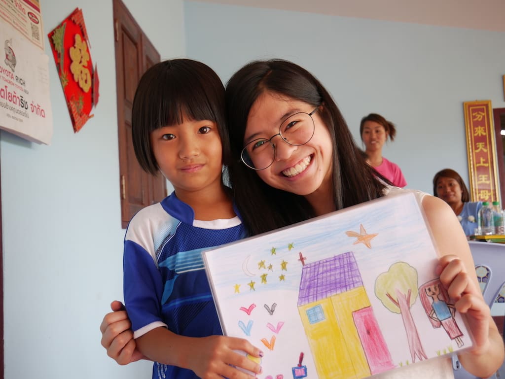 A young woman and a young girl stand together, holding up a child's drawing. Both are looking at the camera, smiling.
