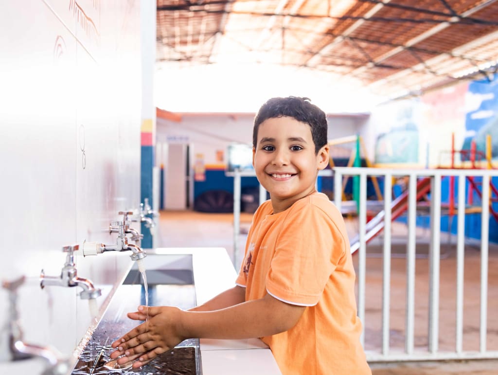 A boy in a orange shirt stands at a sink washing his hands. He is smiling.