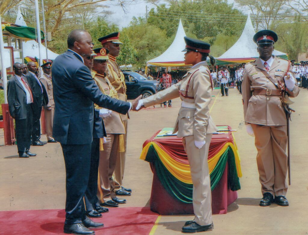 Miriam shakes the hand of the Kenyan president as she receives an award for being the fastest cadet to complete the quick march routine. She is wearing a dress uniform, tan pants and shirt, and black hat. There is a table with red, yellow and green tablecloths behind her.