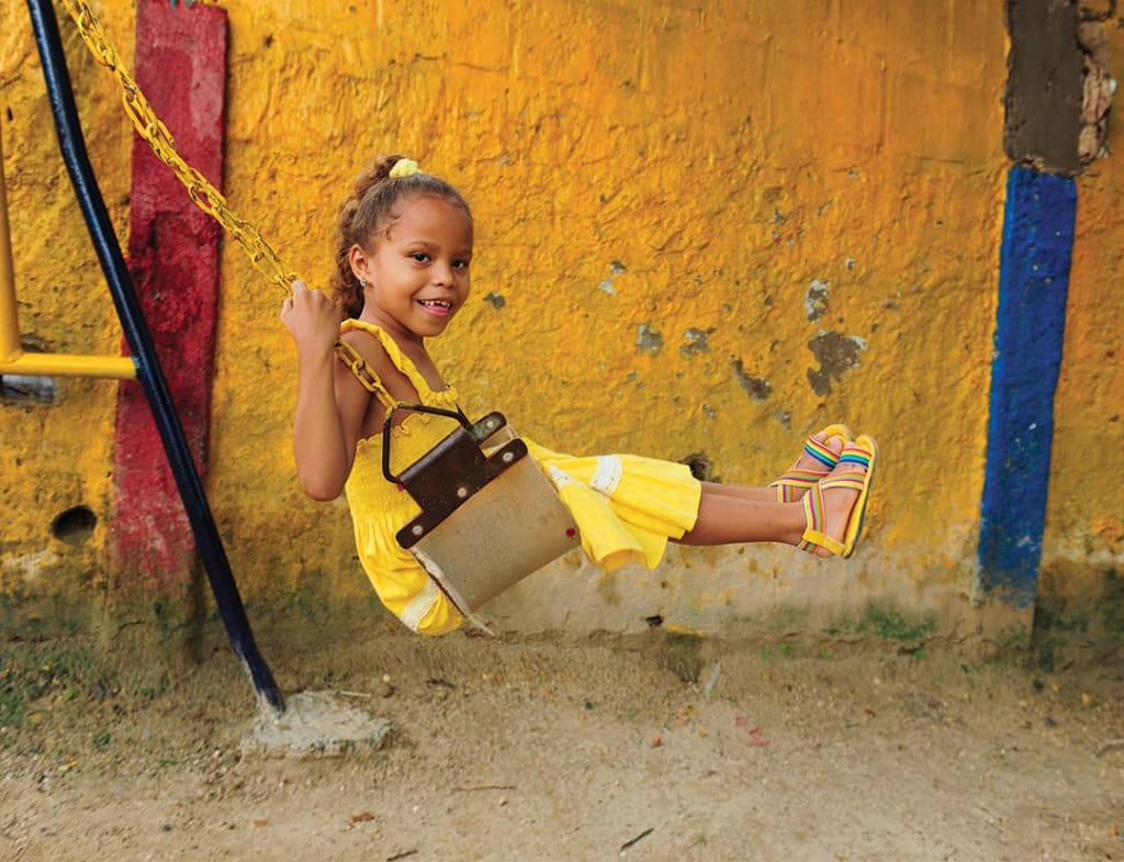 A young girl in a yellow dress, swings on a swing-set and smiles.