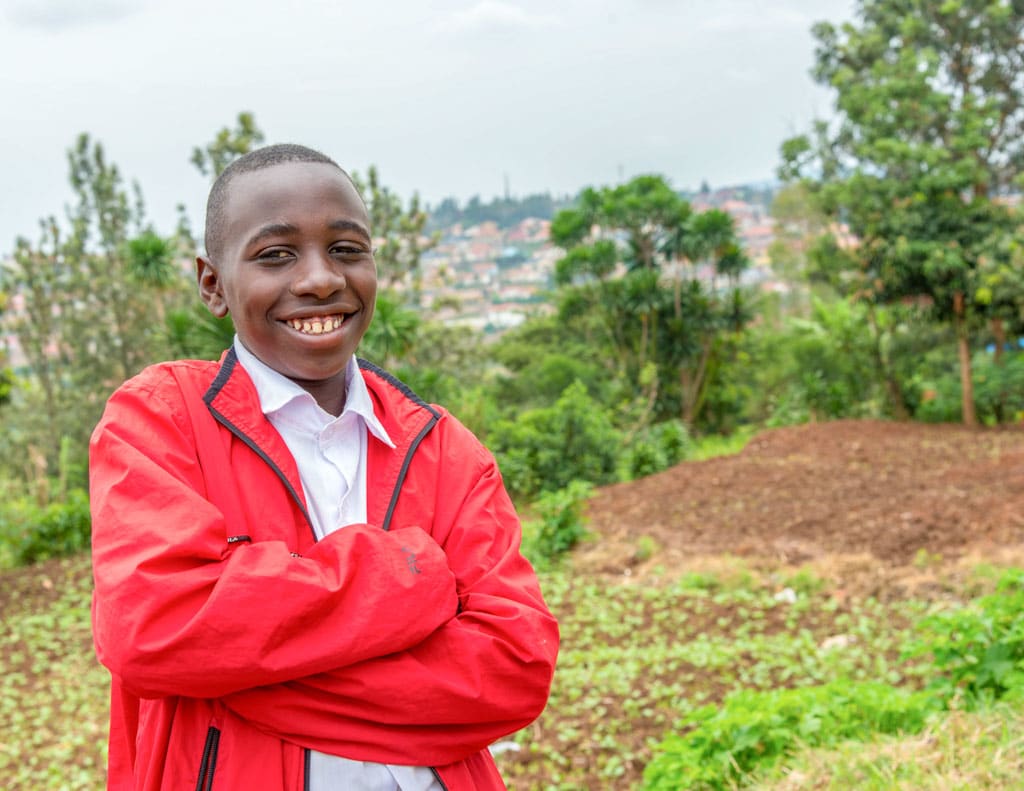 A Rwandan boy wearing bright red jacket, stands with his arms crossed and smiles at the camera.