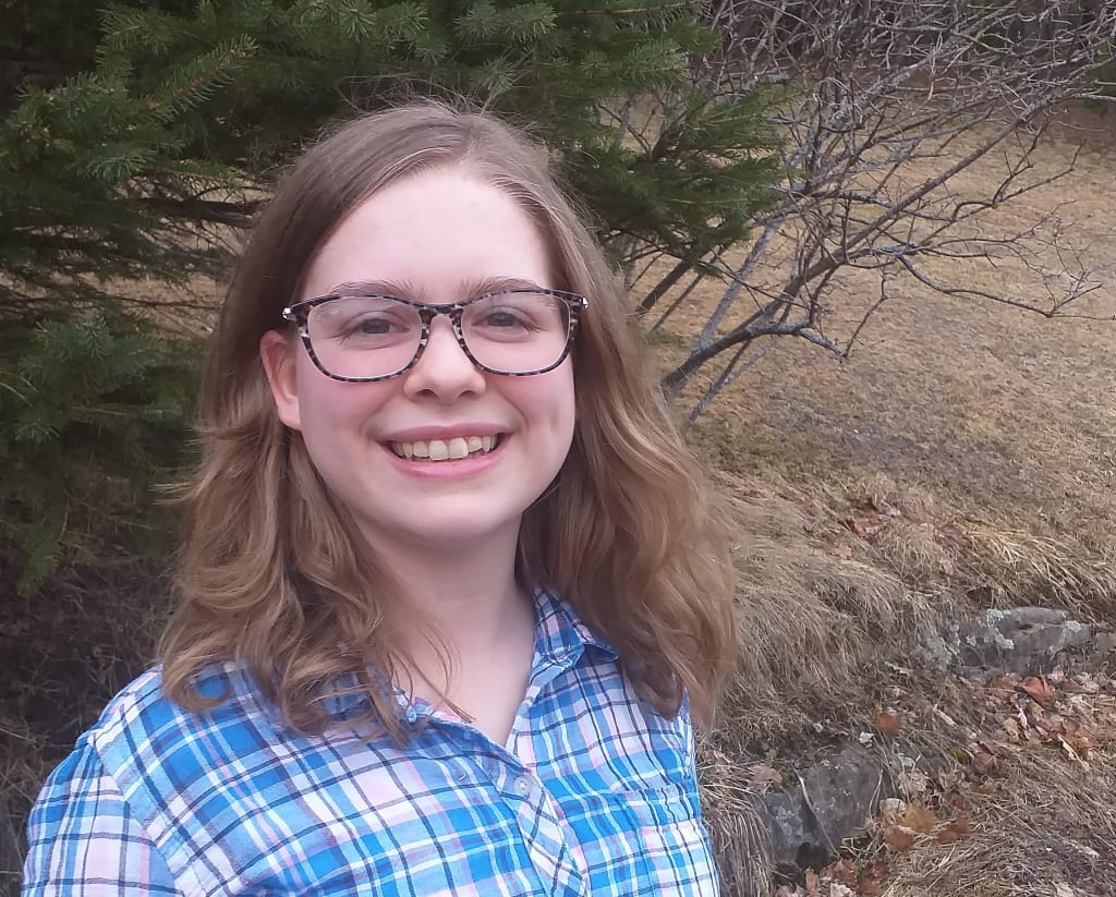 A girl in a blue checked top and glasses smiles at the camera