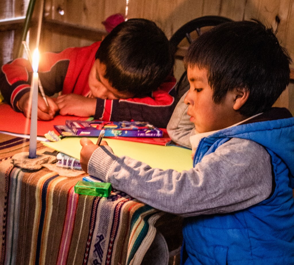 Little boy wearing a blue vest does his homework by candlelight beside his brother wearing a red sweater.