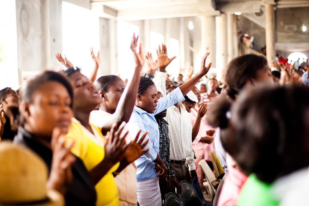 Women stand in an open air church building. They are worshiping with their eyes closed and their hands raised. They are surrounded by many other congregants.