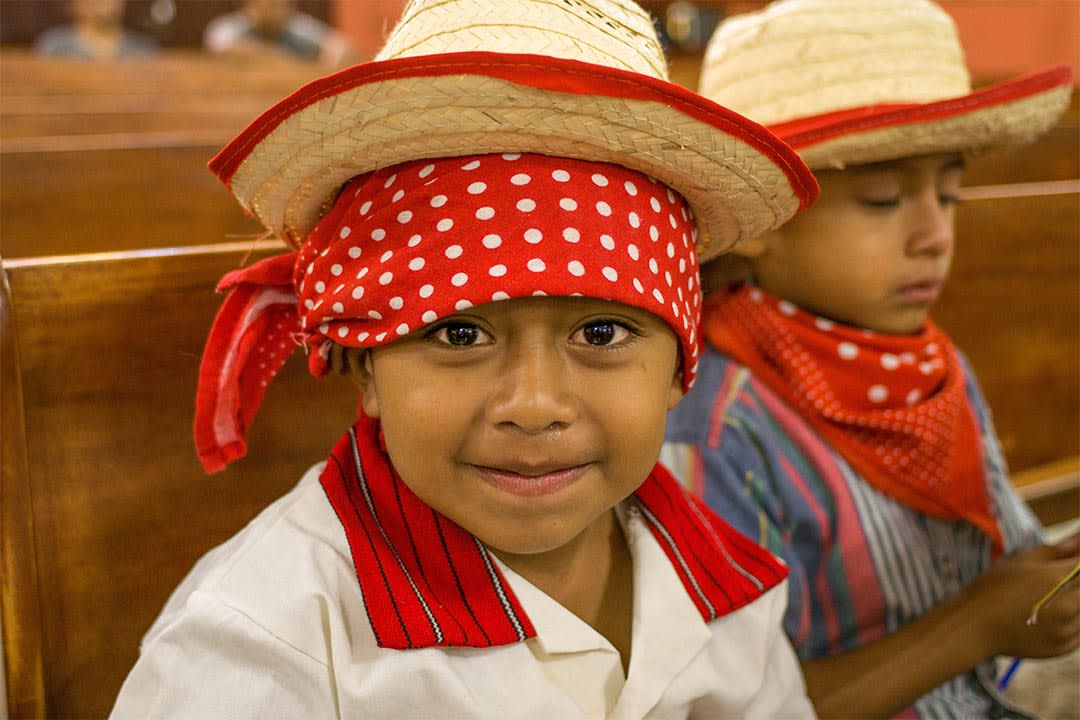 A young boy from Guatemala sits in a church pew. He's wearing straw hat on top of a red bandana. His white shirt has a striped, red collar.