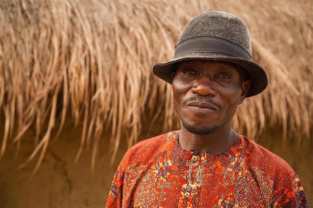 A man from Ghana stands in front of a mud hut with a grass roof. He where's a vibrantly patterned button up shirt and brown dress hat.