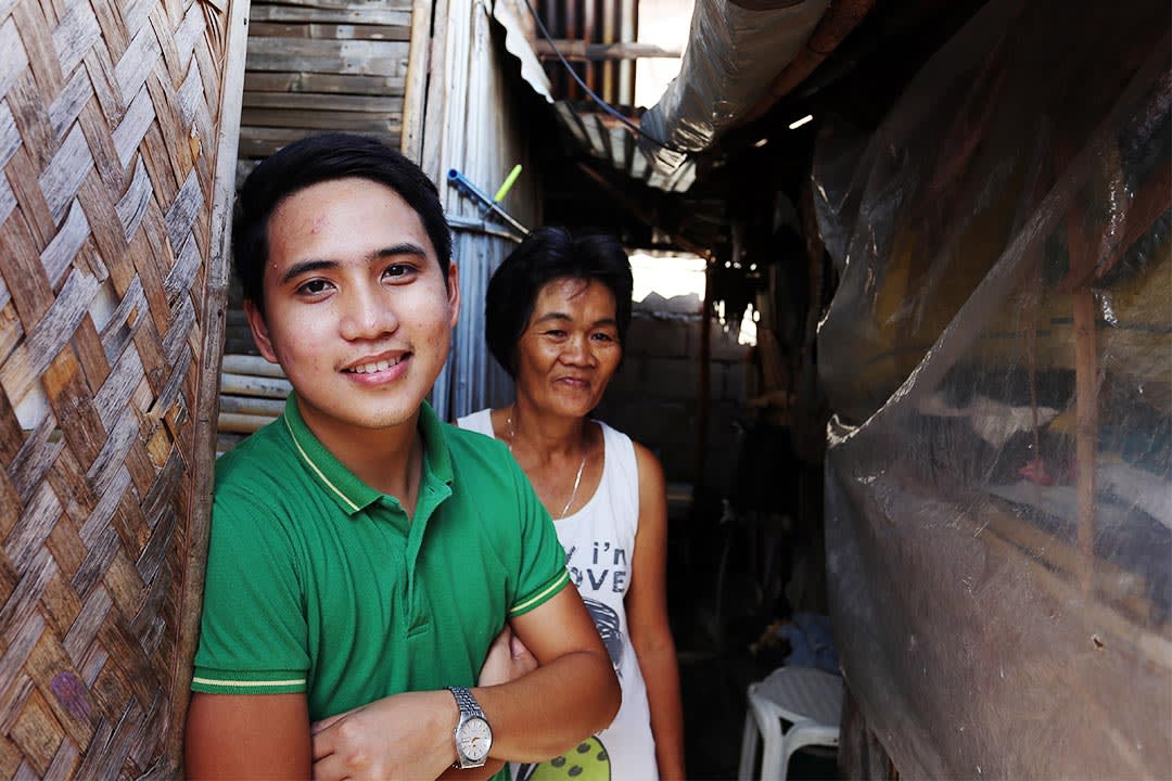 Jeric and his mother stand in front of their home in the Philippines. They are both smiling at the camera
