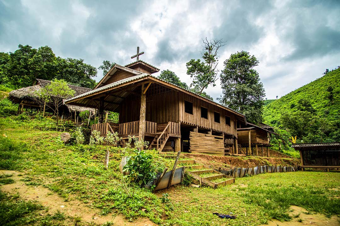 A church in Thailand stands on the side of a hill amidst lush green grass and trees. It is made out of wood and stands above the ground on wooden stilts.