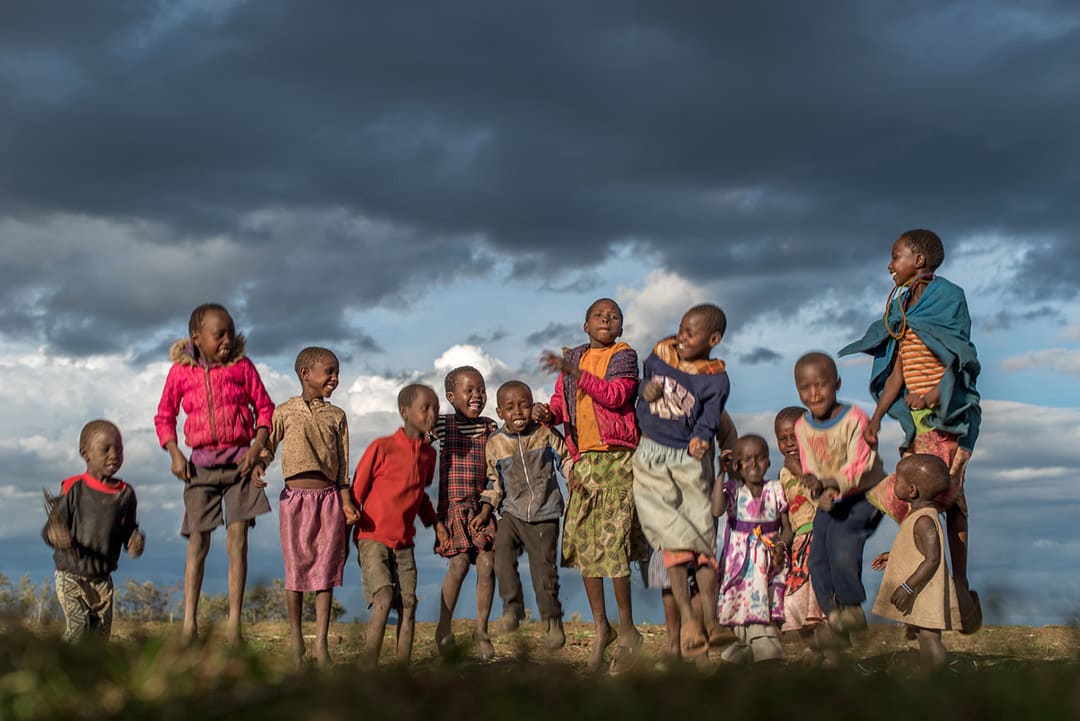 Boys and girls in bright coloured jackets and clothing jump and smile. The sky behind them is cloudy.