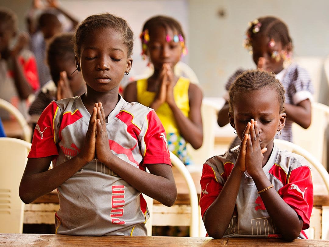 Two girls sit at a desk with their hands folded and their eyes closed. They are praying. They're wearing matching red and grey puma athletic t-shirts.