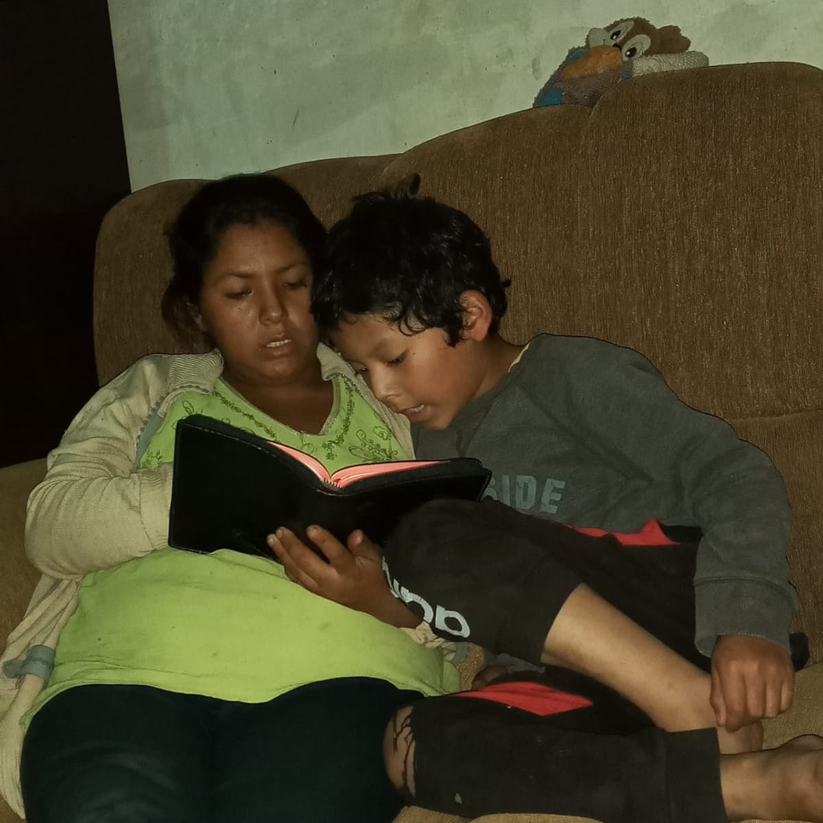 A Bolibian woman sits on a bed with her son and reads the Bible.