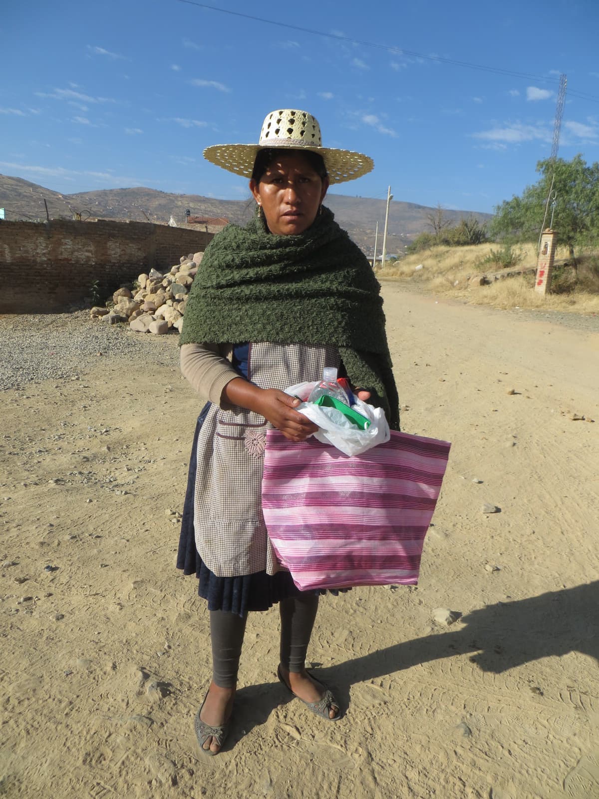 A Bolivan woman, wearing a traditional hat, holds a bag containing a hygiene kit. She stands outside.