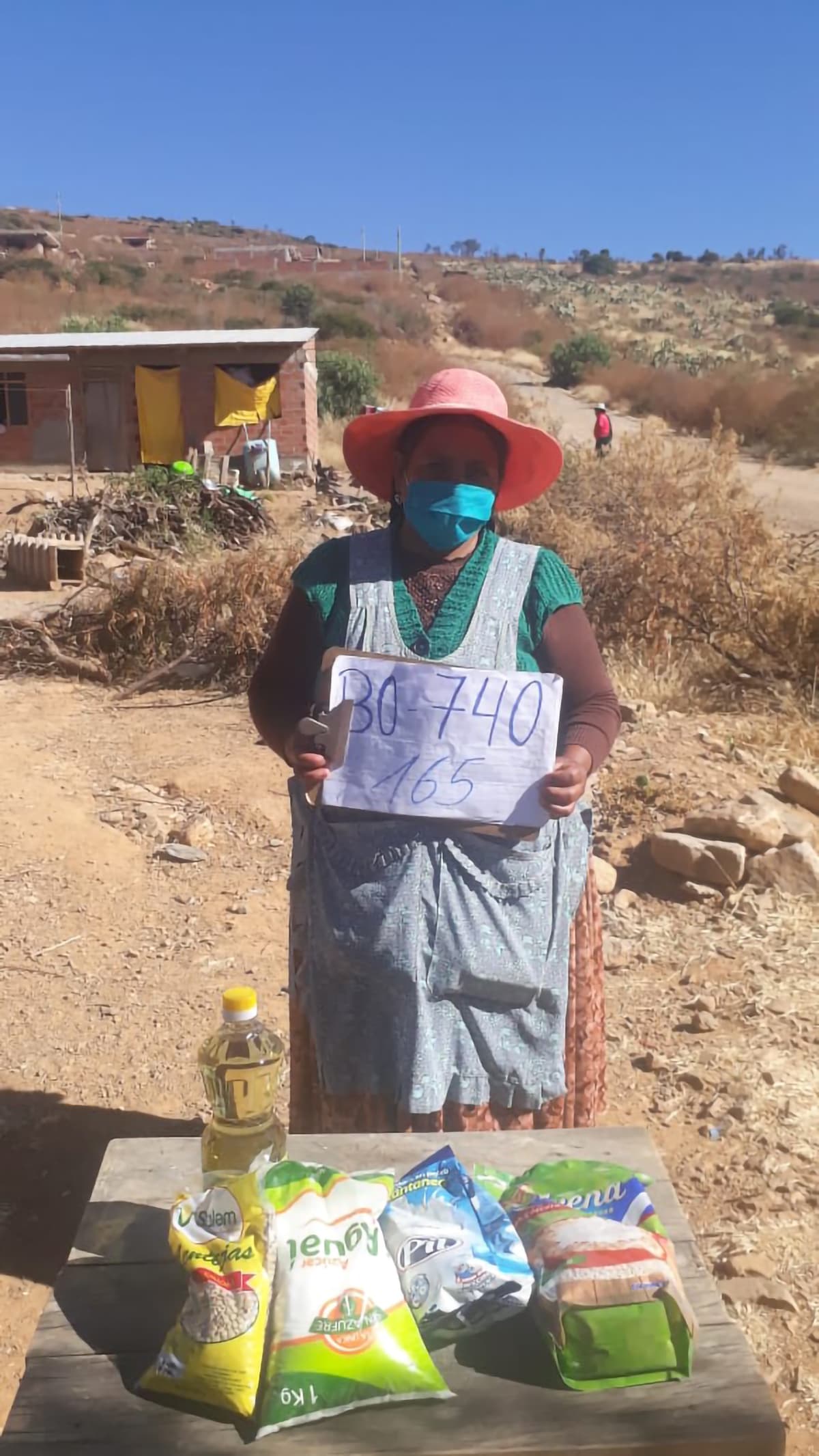 A Bolivian woman stands out side. She wears a surgical mask and stands behind hygiene kit layed out on the table.