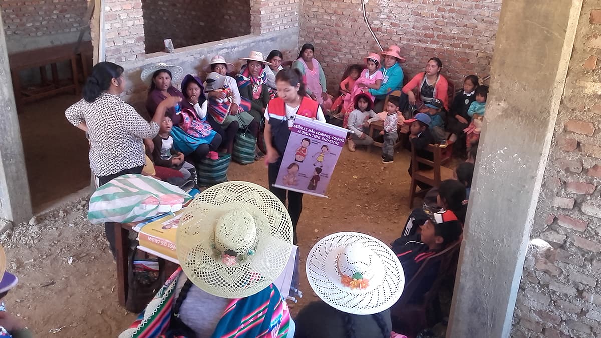 A group of moms sit in circle in a cinder block building and listen to center volunteers teach. Many wear traditional Bolivian garb.