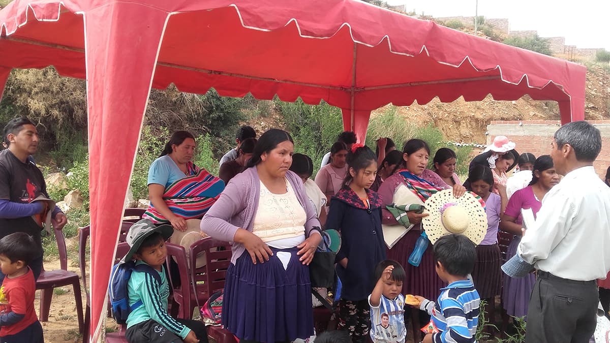 Women men and children sit and stand under red, temporary awning, set up for an outreach program. A man stands in front of the them and speaks.