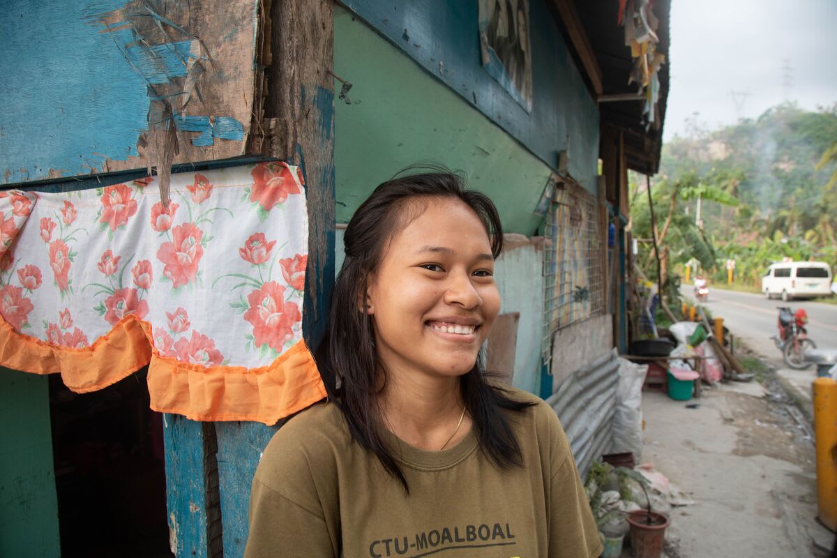 A girl in a brown shirt smiles at the camera in front of a blue and green building