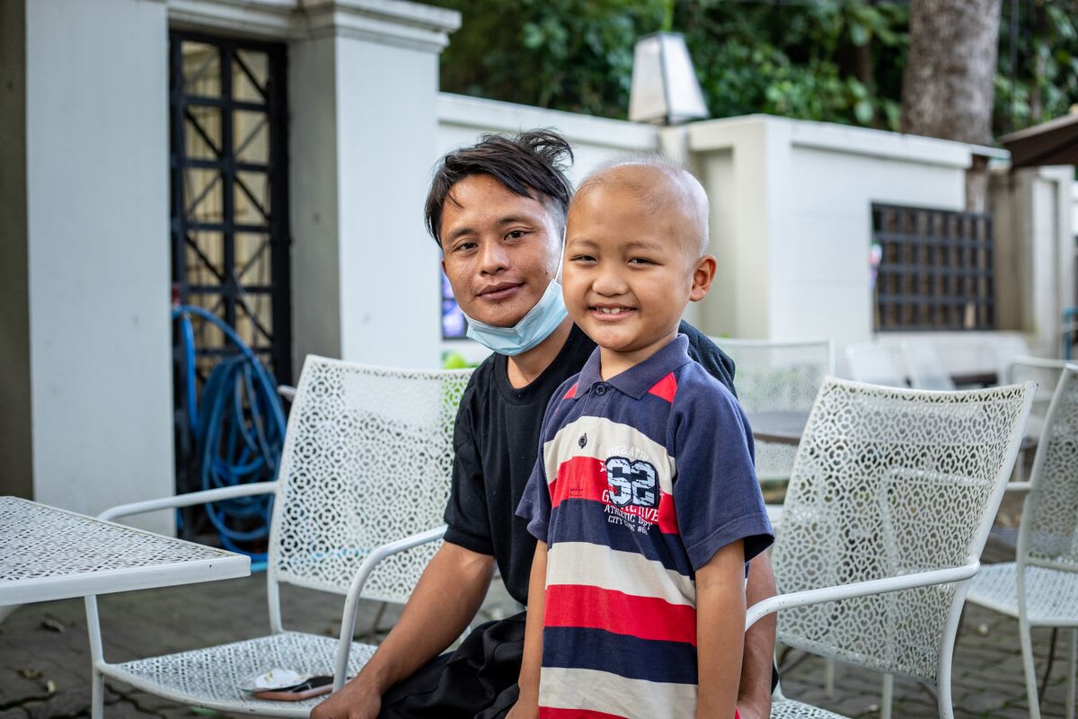 A boy in a blue, red and white striped shirt smiles at the camera with his father smiling behind him