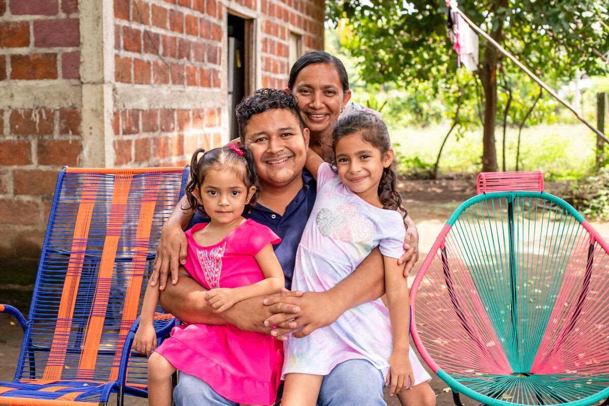 A father sits with his two young daughters on his lap and his wife hugging them from behind. They are sitting in between two colourful chairs and they are smiling