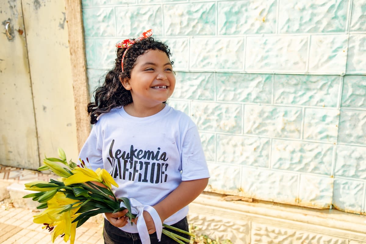 Valery is smiling as she holds a bouquet of flowers while standing outside in front of a building in the community. She is wearing a t-shirt that says “Leukemia Warrior” on it.
