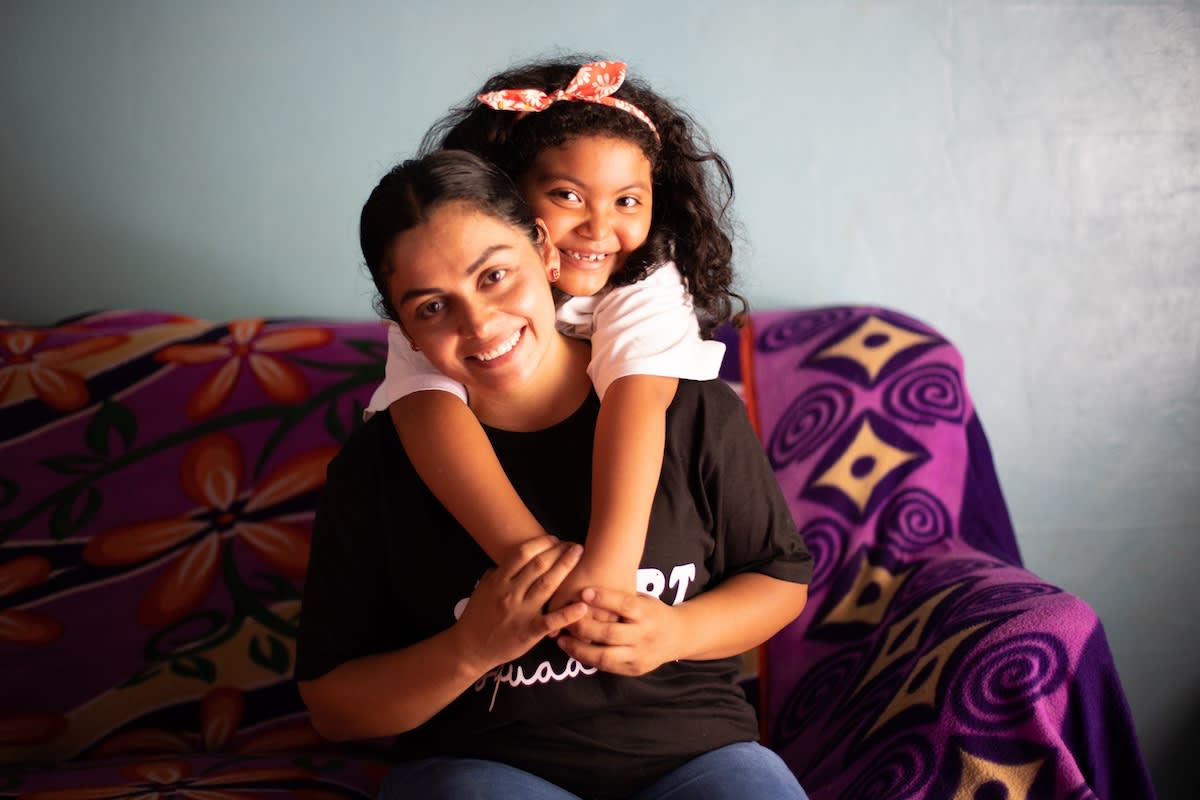 Portrait of Valery and Yesenia at home. Yesenia is sitting on the couch and Valery is behind her hugging her. They are both smiling.