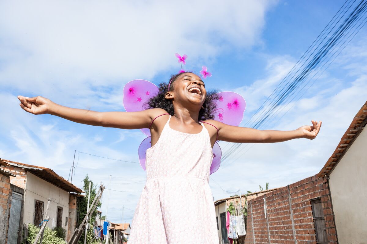 A girl in a pink dress and pink butterfly wings laughs while holding her arms stretched wide under a blue sky