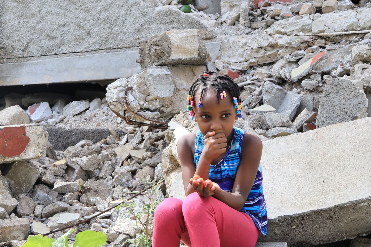 A girl in a blue top and pink pants sits in a pile of rubble and has her hand at her mouth.