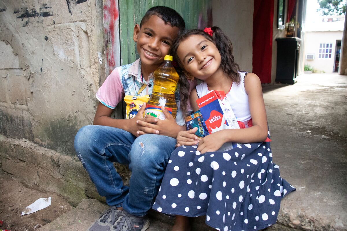 Luis is wearing a blue and white patterned shirt with pink sleeves and jeans. He is sitting outside his home with his sister, María Jose, wearing a blue and white dress. They are each holding food supplies from the centre.