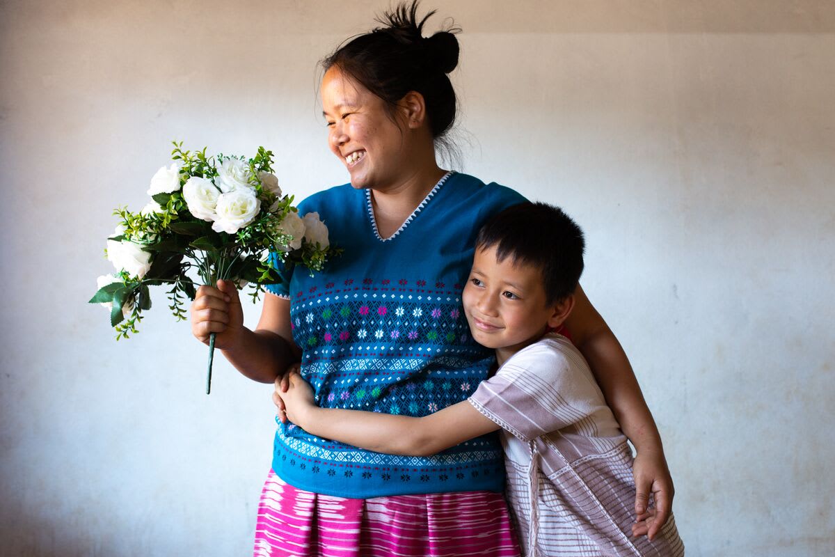 A small boy is wearing a white shirt. He is with his mother who is wearing a blue shirt and a pink skirt. He has his arms around his mother and she is holding a bouquet of white flowers.