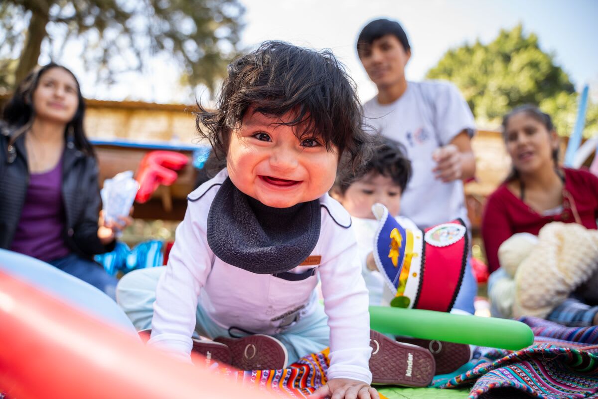 A baby is wearing a white shirt and a black bib. He is smiling at the camera. He is playing outside and is surrounded by survival program mothers.
