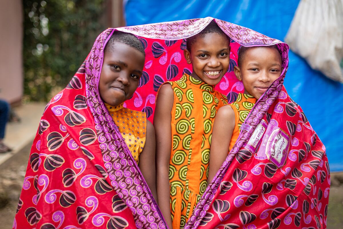 Three girls in orange dresses smile at the camera while wrapped up in a bright pink fabric