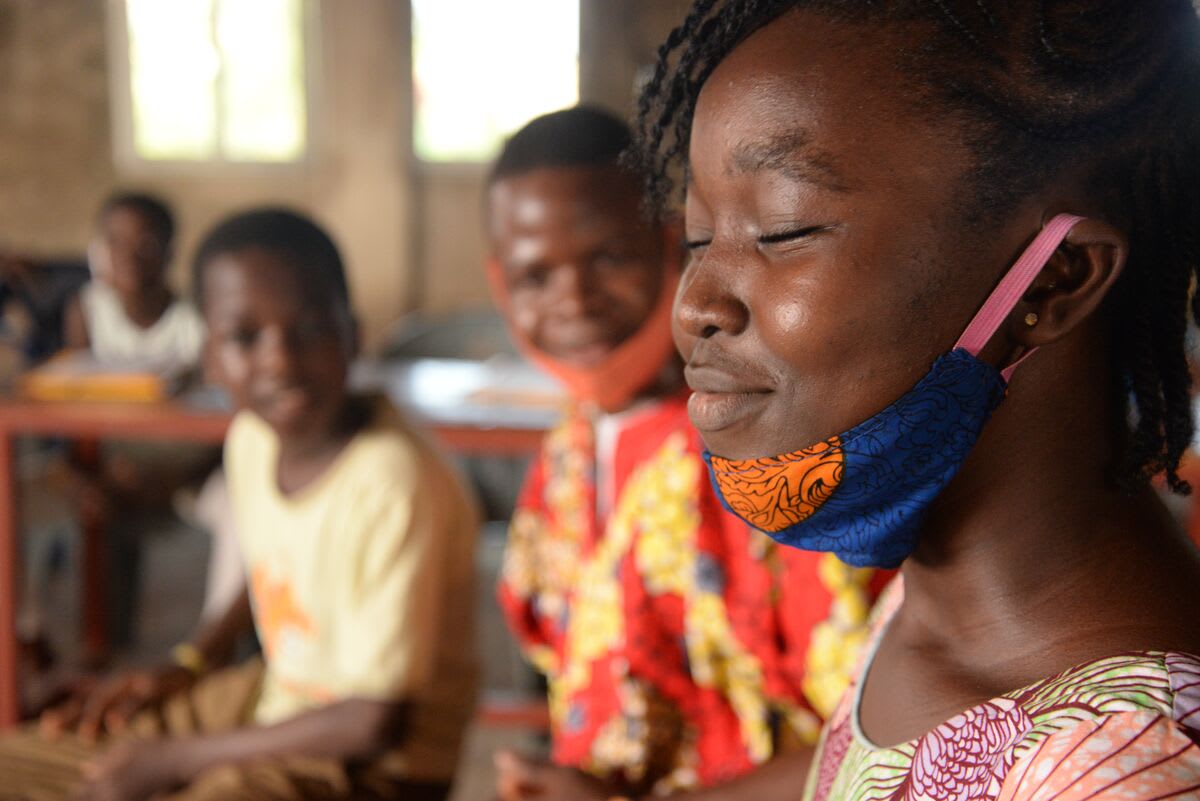 Olivia is wearing a pink and green floral print dress with a blue face mask under her chin. She is acting in a skit. There are other beneficiary children in the background.