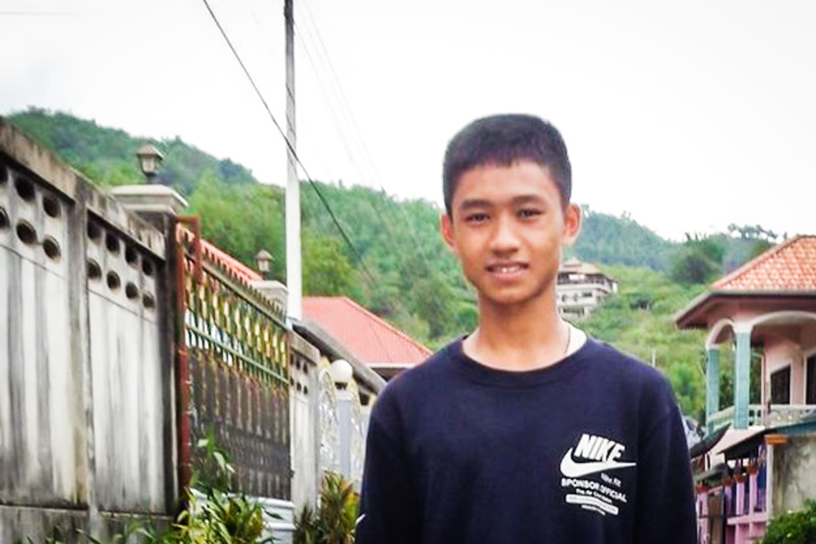 A boy is smiling, wearing a black Nike shirt. Behind him is a fence and a hill of green trees.