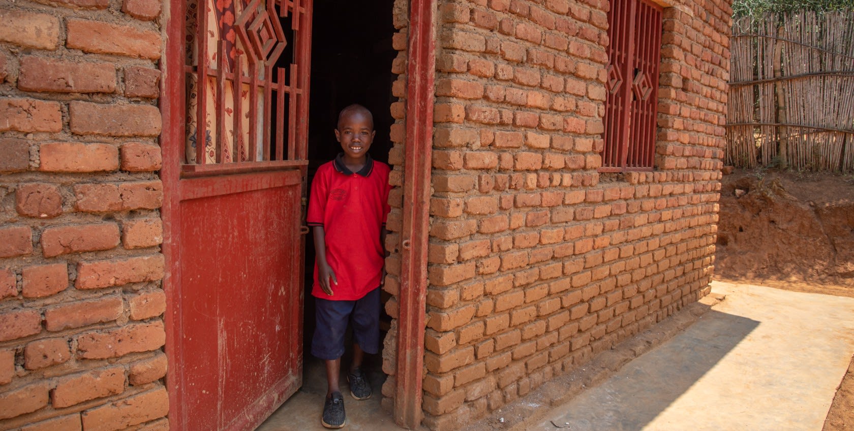 A boy wearing red shirt, standing by the door of a brick house.