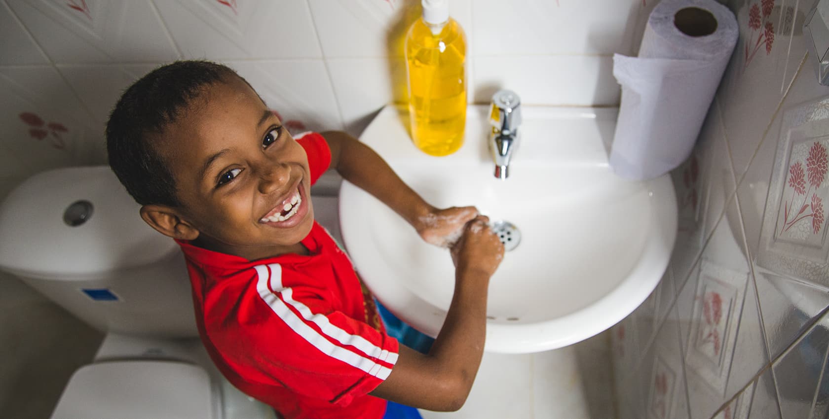 A boy in a red t-shirt washes his hands in sink in a bathroom. He's missing a front tooth.