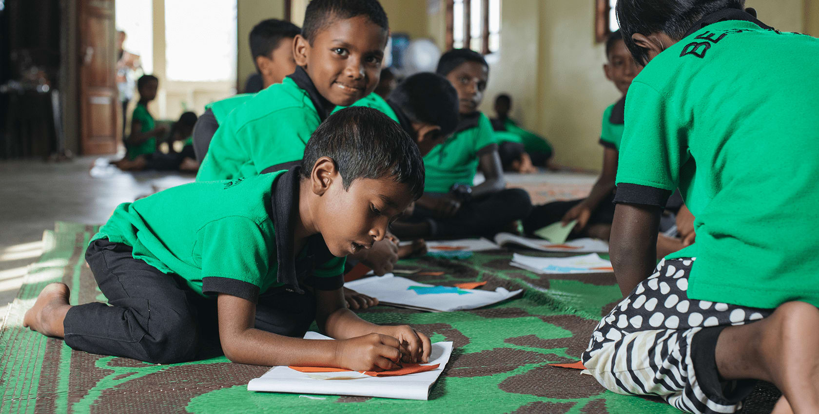 Boys wearing green school uniforms sit at desks and write on work sheets
