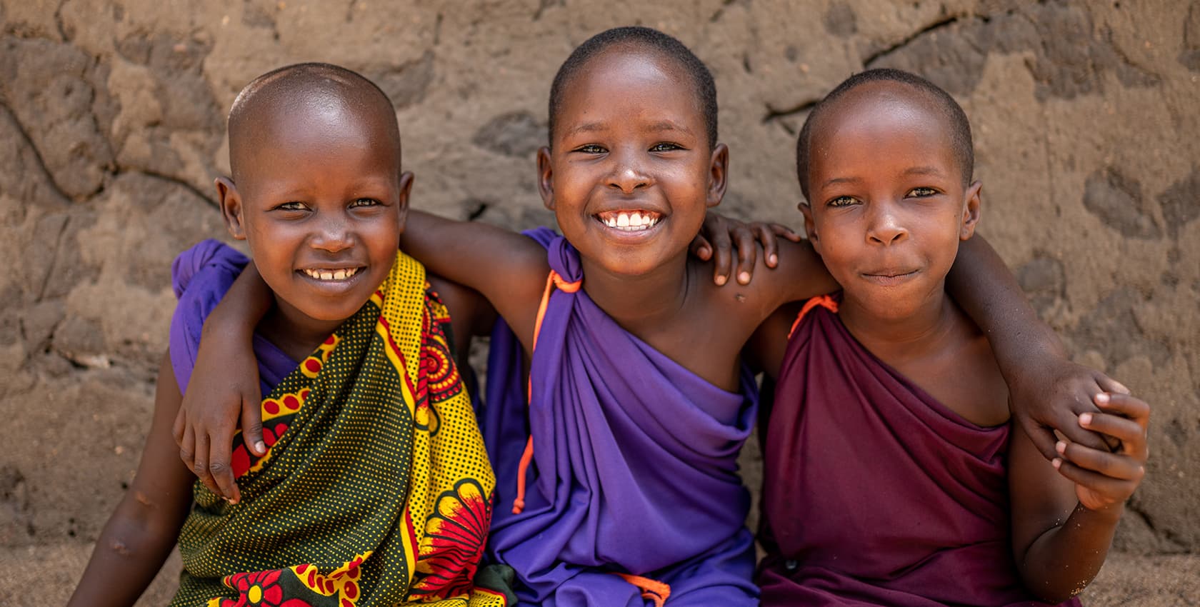 Three grade school age girls sit together in front of a stone wall and smile.