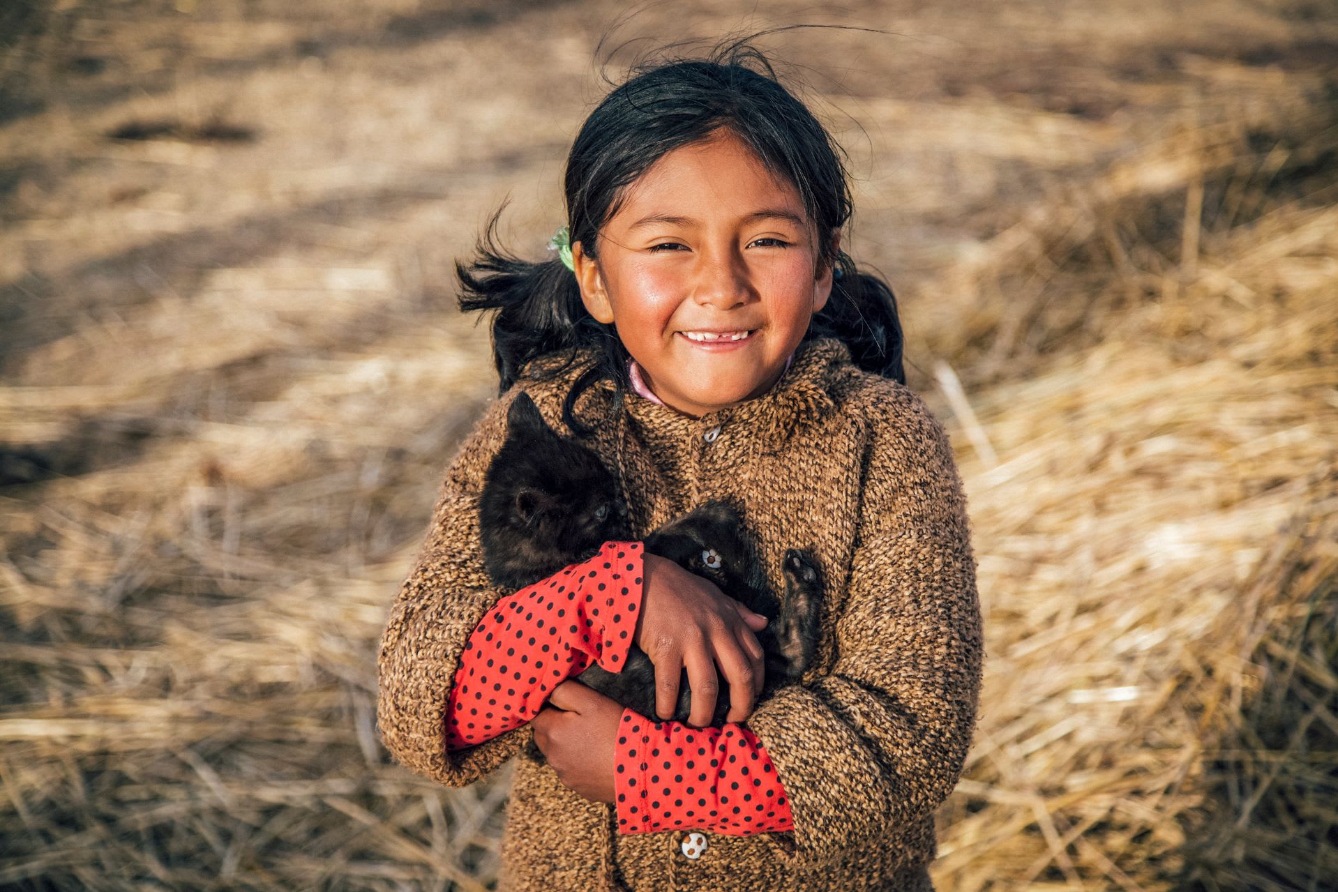 A girl in a brown sweater holds a black kitten while smiling.