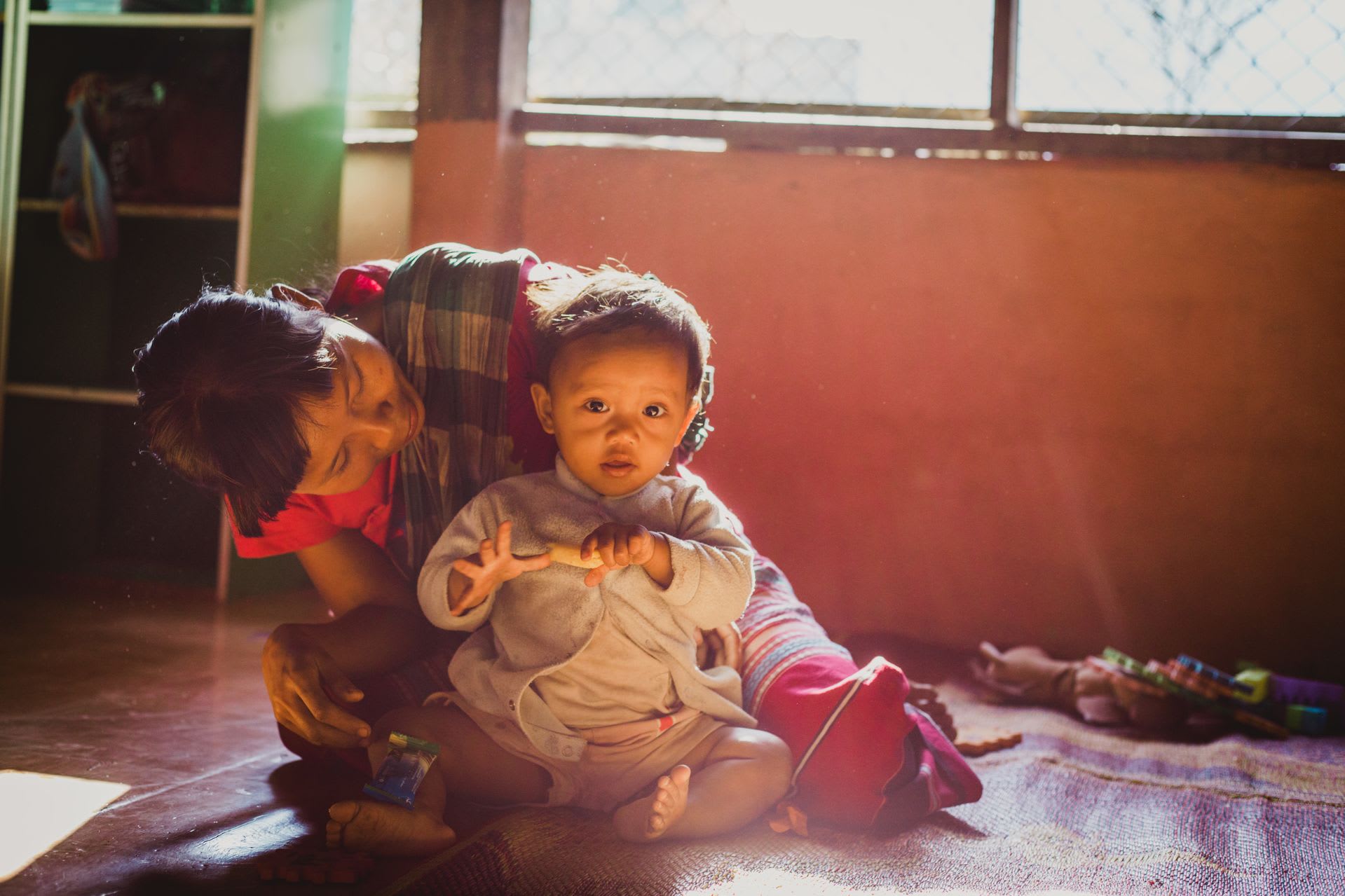 Patanree sits cross legged on the ground with her toys, looking at the camera as the sun beams into their hoe. Her mom sits behind her smiling at her.