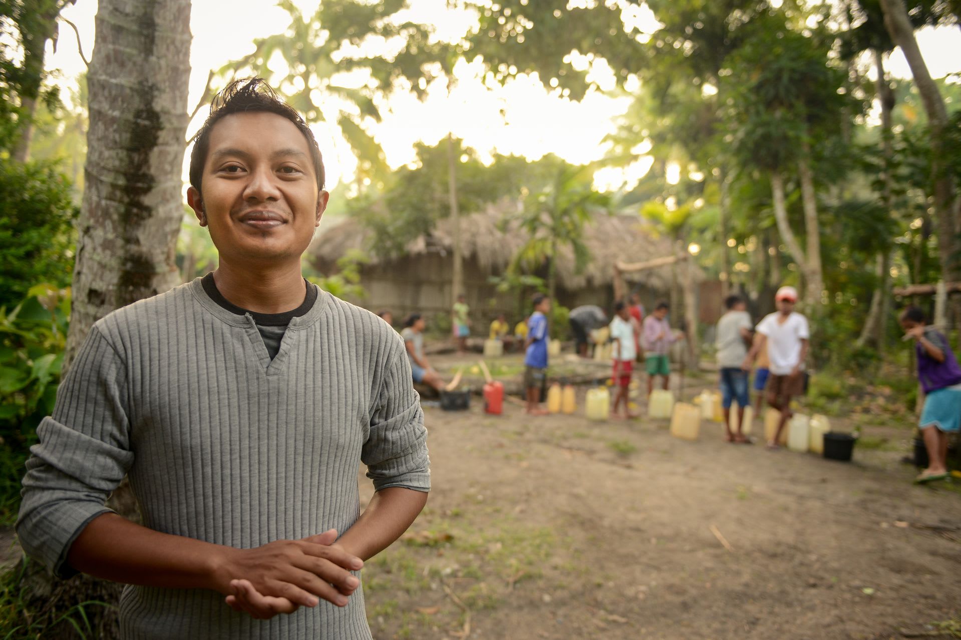A man in a gray shirt stands outside in front of palm trees and a grass thatched building at sunset with people in a line in the background gathering water