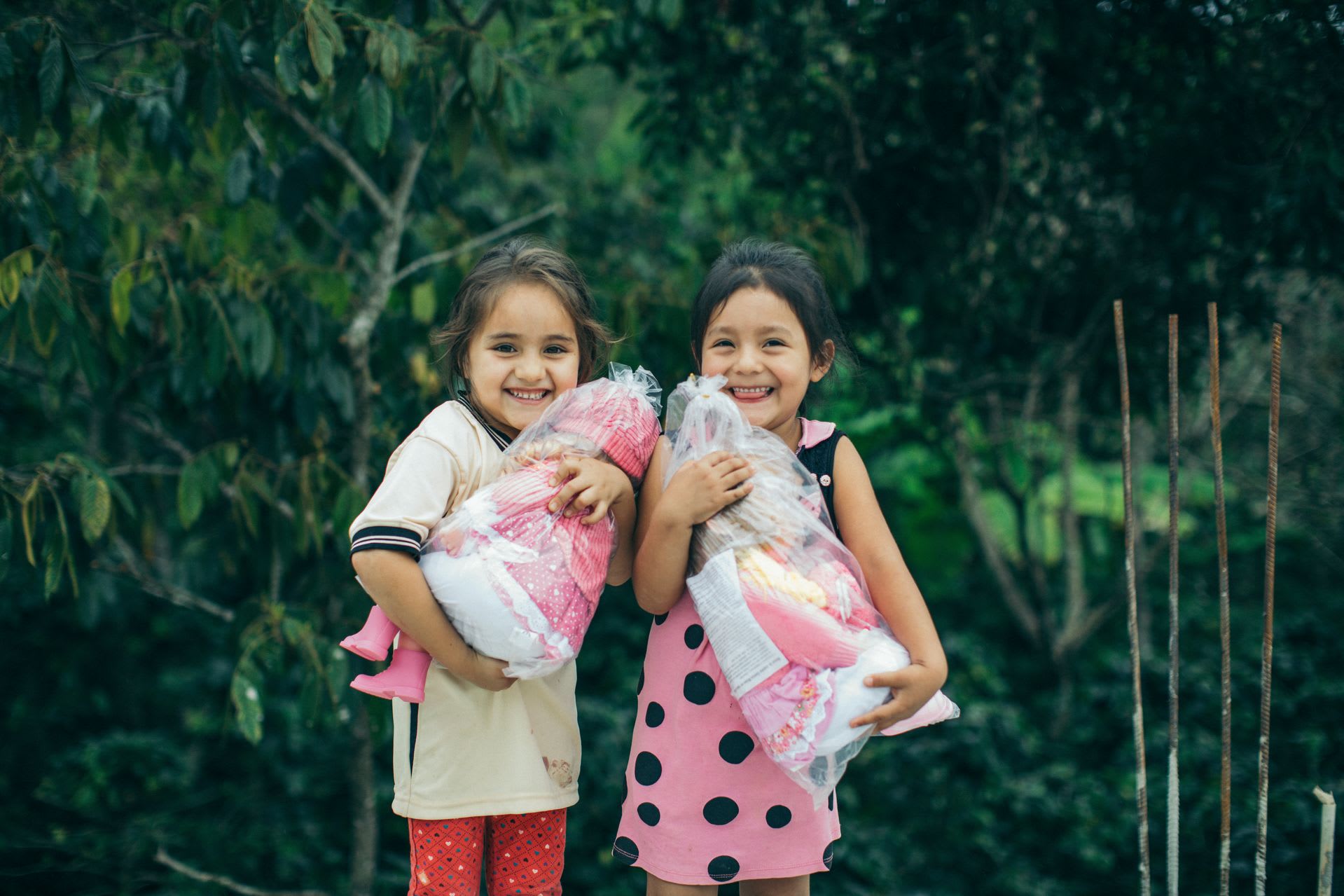 Two little girls are holding new dolls. They are smiling big with trees in the background.