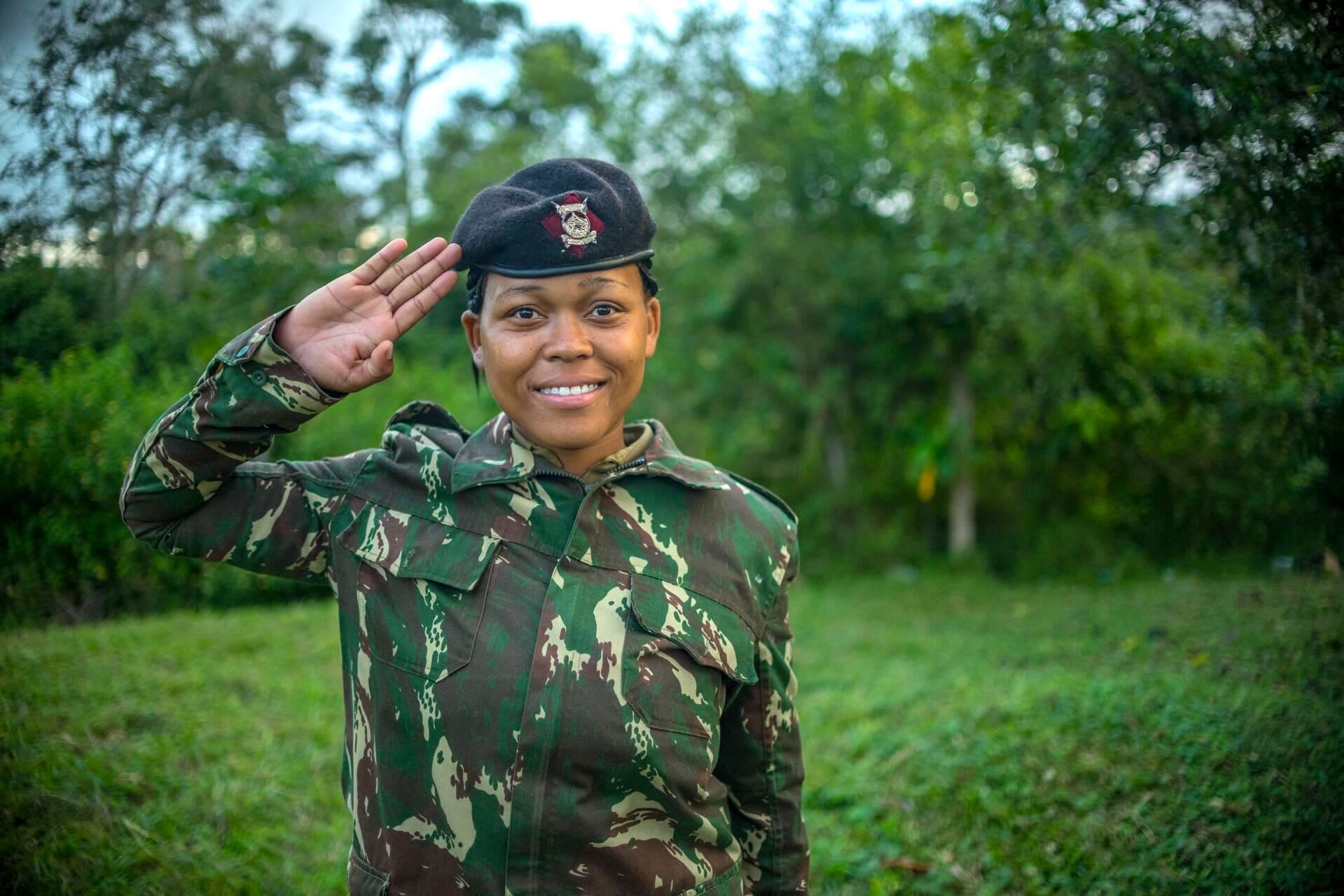 Miriam is pictured here in her uniform - a camoflauge top and green pants. She is wearing a hat. She is smilimg at the camera and saluting, and there are trees in the background.