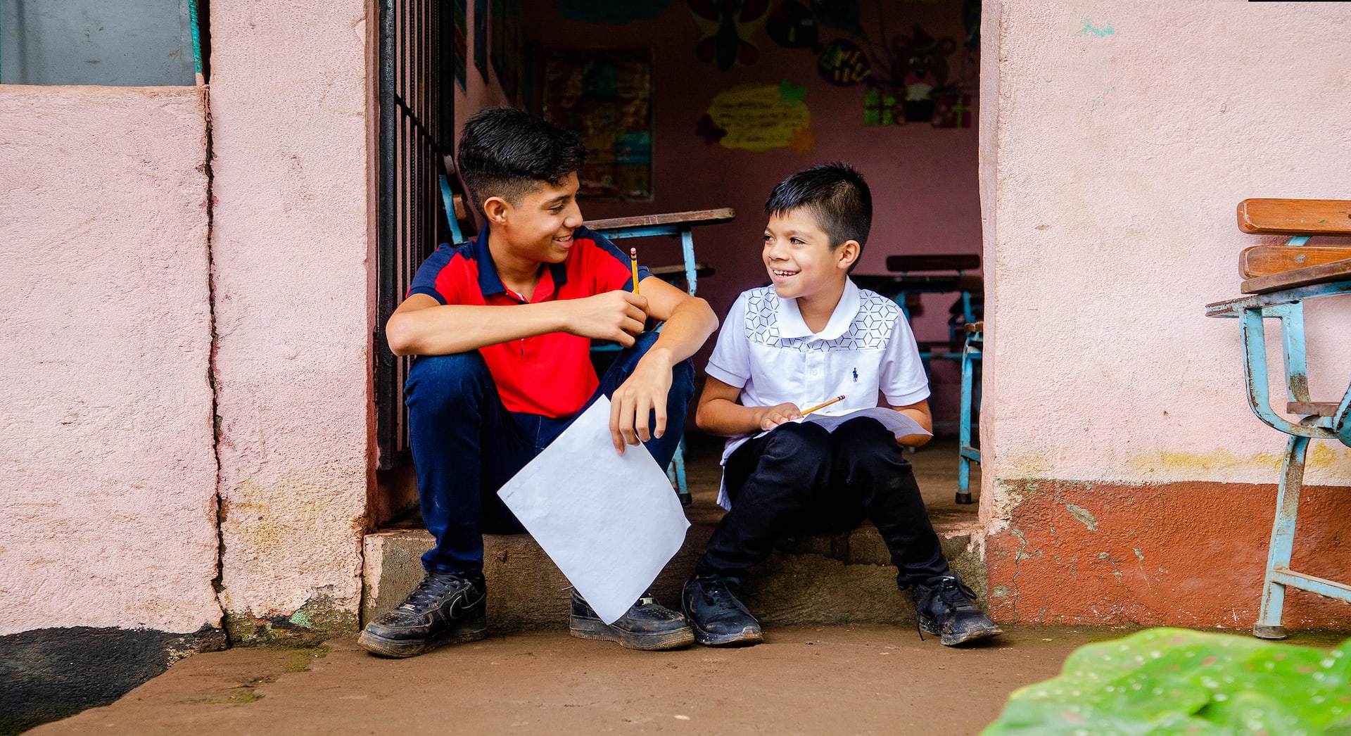 Two young boys sit in the doorway on their home holding a letter and smiling at each other.