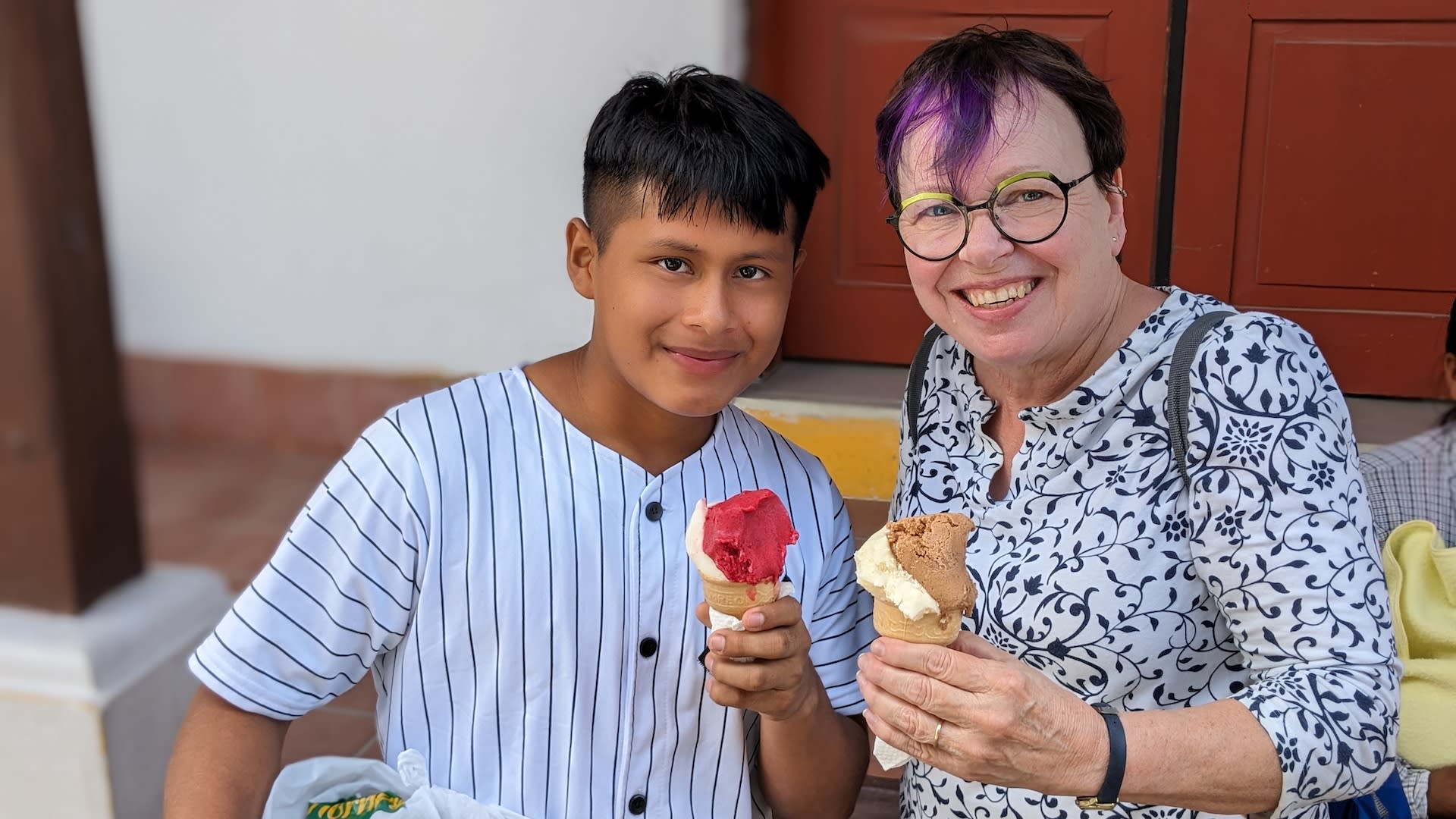 Lynn is wearing glasses and a floral shirt. She holds an ice cream cone. Roberto is wearing a striped shirt and holding an ice cream cone.